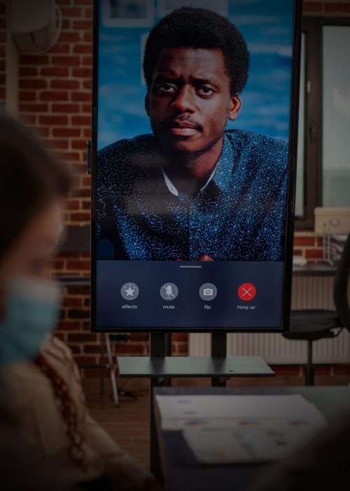 Black man seen on led screen panel, conducts videoconference call with team in brick wall office. Digital monitor shows ongoing virtual meeting of client, representing remote communication in office.